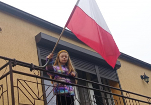 Uczennica stoi na balkonie i trzyma flagę biało-czerwoną.